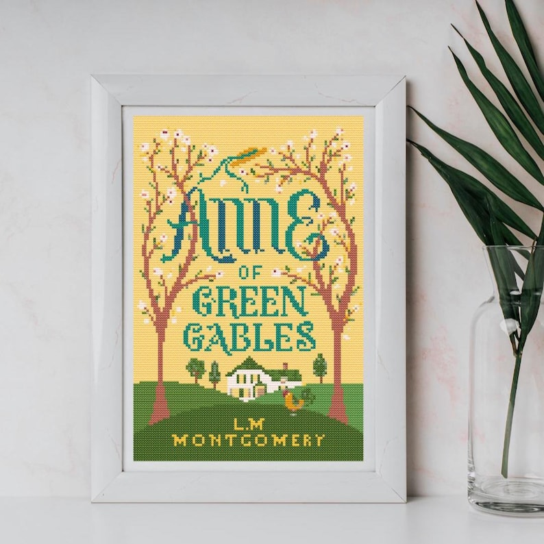May include: Framed cross-stitch art featuring the book cover of "Anne of Green Gables." The design includes the title in teal and white, with a yellow background. The artwork depicts trees, a house, and the author's name.