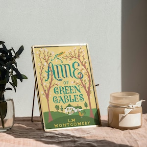 May include: A framed cross-stitch of "Anne of Green Gables" by L.M. Montgomery. The design features the title in teal, a house, and trees on a yellow background. The frame is gold, and the piece is propped up on a stand. A candle and plant are nearby.