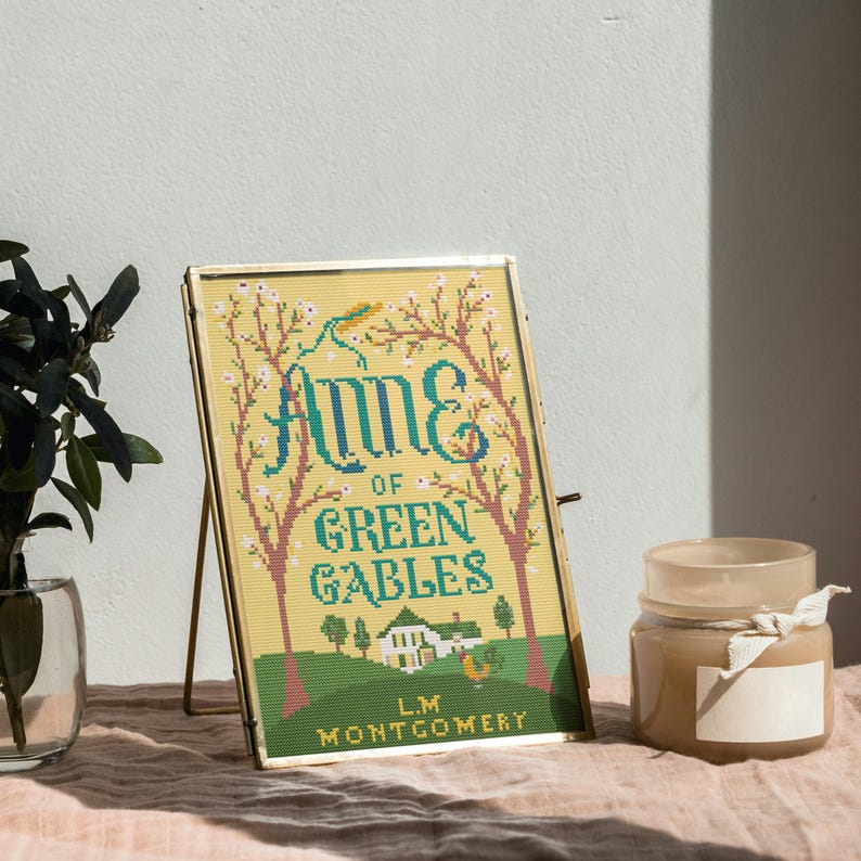 May include: A framed cross-stitch of "Anne of Green Gables" by L.M. Montgomery. The design features the title in teal, a house, and trees on a yellow background. The frame is gold, and the piece is propped up on a stand. A candle and plant are nearby.