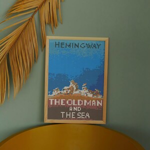 Könnte beinhalten: Ein gerahmtes Kreuzstich-Kunstwerk mit einem blauen und weißen pixelartigen Bild eines Bootes auf dem Meer. Der Text "Hemingway" befindet sich oben und "Der alte Mann und das Meer" unten.