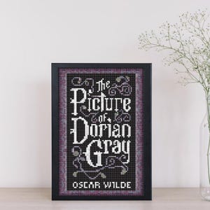 May include: A black framed cross-stitch of the book cover for "The Picture of Dorian Gray" by Oscar Wilde. The title is in white with a purple and gray background.