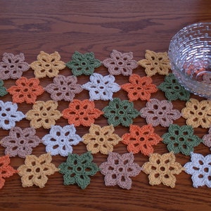 May include: A crocheted table runner with a pattern of flowers in shades of orange, green, brown, white, and yellow. The runner is laid out on a wooden table with a glass bowl on top.