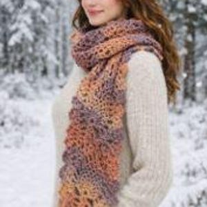 Puede incluir: Una bufanda larga de ganchillo con un patrón de colores variados en tonos naranja, morado y beige. La bufanda se lleva sobre un suéter de punto de color crema de manga larga con textura acanalada, creando un estilo de capas para el frío.
