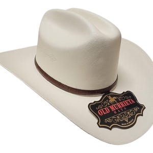 Puede incluir: Sombrero de vaquero blanco roto con una banda de cuero marr&oacute;n y un ala curvada. El sombrero presenta la etiqueta "OLD MURRIETA HATS". Este sombrero de estilo western es adecuado para uso casual y formal.