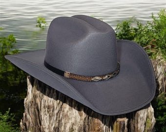 Puede incluir: Un sombrero vaquero de fieltro gris con una banda de cuero marrón y detalles plateados. El sombrero está sentado en un tocón de árbol.