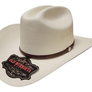 Puede incluir: Sombrero de vaquero blanco roto con una banda de cuero marr&oacute;n y una etiqueta decorativa. El sombrero tiene un dise&ntilde;o cl&aacute;sico con ala curva y copa estructurada. La etiqueta dice "OLD MURRIETA HATS".