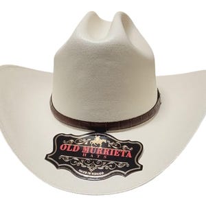 Puede incluir: Sombrero de vaquero color crema con una banda de cuero marr&oacute;n. El sombrero tiene un ala ancha y una corona alta. Una etiqueta negra y roja en el ala dice "OLD MURRIETA HATS" y "MADE IN MEXICO".