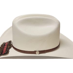 Puede incluir: Un sombrero de vaquero blanco con una banda de cuero marr&oacute;n y una hebilla plateada. El sombrero tiene una forma cl&aacute;sica con un ala ancha y una corona ligeramente hundida. El sombrero tiene una etiqueta decorativa que dice "Old Marietta".