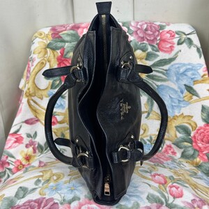 Puede incluir: Bolso de mano de cuero negro con cremallera superior y dos asas. El bolso tiene herrajes plateados y un peque&ntilde;o logotipo en la parte delantera. El interior es visible y el bolso est&aacute; abierto. El bolso est&aacute; sobre una superficie con estampado floral.