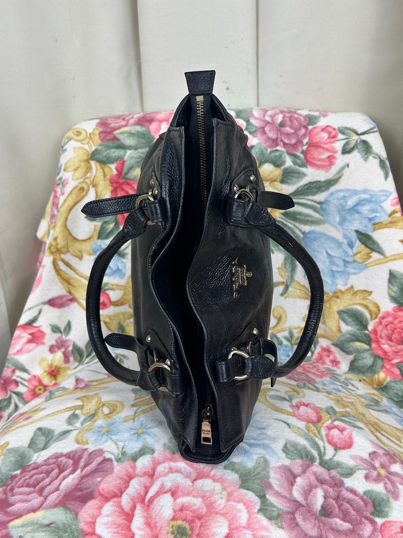 Puede incluir: Bolso de mano de cuero negro con cremallera superior y dos asas. El bolso tiene herrajes plateados y un peque&ntilde;o logotipo en la parte delantera. El interior es visible y el bolso est&aacute; abierto. El bolso est&aacute; sobre una superficie con estampado floral.