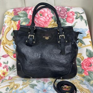Puede incluir: Bolso tote Prada de cuero negro con asas dobles y una correa de hombro desmontable. El bolso presenta herrajes plateados, incluyendo hebillas y tachuelas. El logotipo de Prada es visible en la parte delantera.