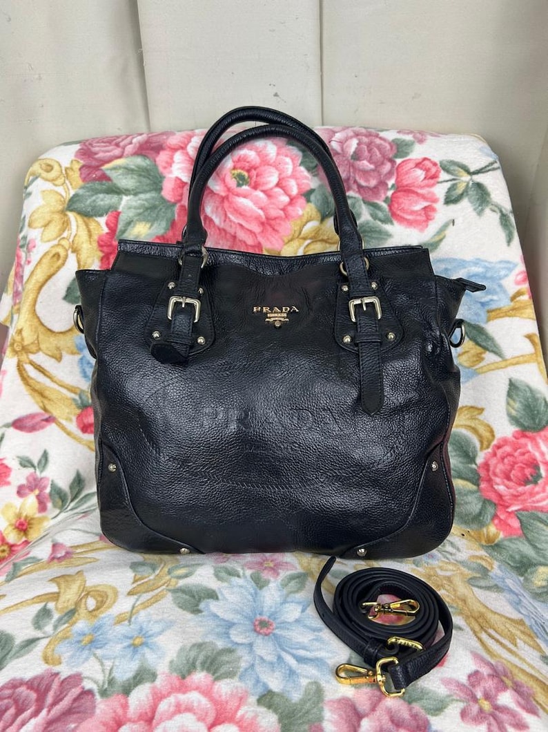 Puede incluir: Bolso tote Prada de cuero negro con asas dobles y una correa de hombro desmontable. El bolso presenta herrajes plateados, incluyendo hebillas y tachuelas. El logotipo de Prada es visible en la parte delantera.