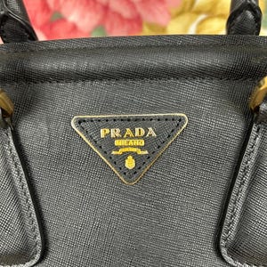 Op de afbeelding: Zwarte leren handtas met een gouden Prada Milano logo. De tas heeft een gestructureerd ontwerp met handvatten aan de bovenkant en een driehoekig logo. De achtergrond is een vage bloemenprint.