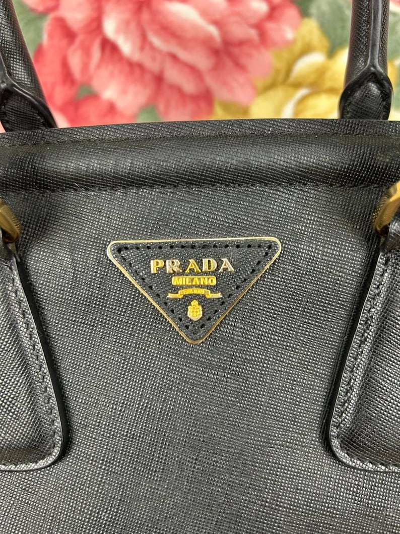 Op de afbeelding: Zwarte leren handtas met een gouden Prada Milano logo. De tas heeft een gestructureerd ontwerp met handvatten aan de bovenkant en een driehoekig logo. De achtergrond is een vage bloemenprint.