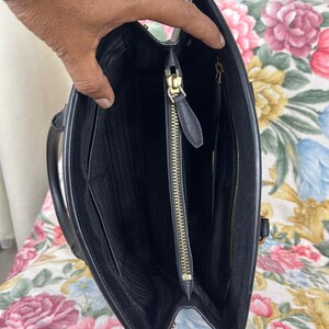 Op de afbeelding: Zwarte leren handtas met een gestructureerd ontwerp. De binnenkant heeft meerdere compartimenten, waaronder een vak met ritssluiting met een goudkleurige ritssluiting. De tas heeft een handvat en een afneembare schouderriem.