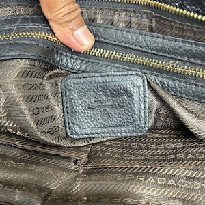 Puede incluir: Primer plano del interior de un bolso de mano de cuero negro. El bolso tiene una cremallera dorada y una etiqueta de cuero texturizado. El forro es de tela marr&oacute;n estampada con el nombre de la marca impreso repetidamente.