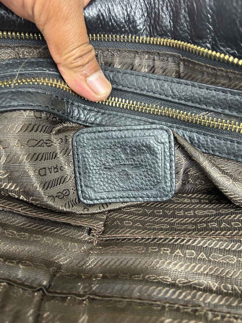 Puede incluir: Primer plano del interior de un bolso de mano de cuero negro. El bolso tiene una cremallera dorada y una etiqueta de cuero texturizado. El forro es de tela marr&oacute;n estampada con el nombre de la marca impreso repetidamente.