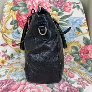 Puede incluir: Un bolso de mano de cuero negro con una superficie texturizada. El bolso tiene dos asas y una correa para el hombro desmontable. Los herrajes met&aacute;licos plateados acent&uacute;an el dise&ntilde;o. El bolso est&aacute; sobre una silla con estampado floral.