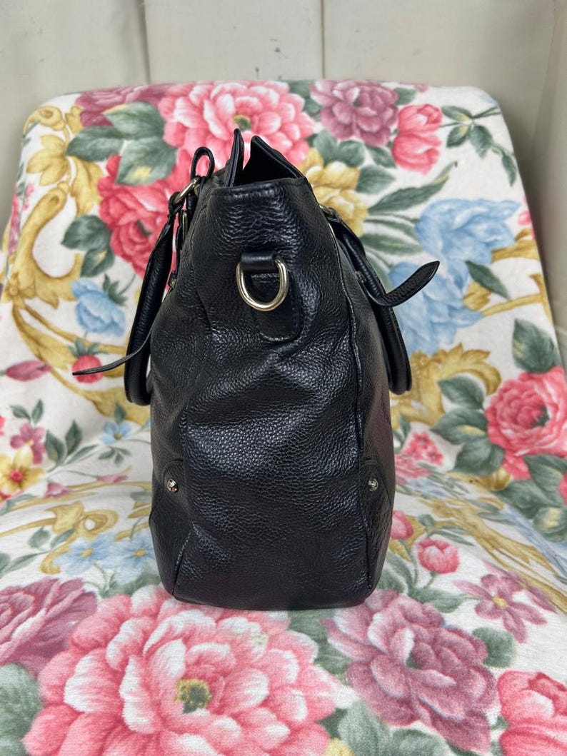 Puede incluir: Un bolso de mano de cuero negro con una superficie texturizada. El bolso tiene dos asas y una correa para el hombro desmontable. Los herrajes met&aacute;licos plateados acent&uacute;an el dise&ntilde;o. El bolso est&aacute; sobre una silla con estampado floral.