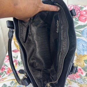 Puede incluir: Bolso de mano de cuero negro con asa superior y cierre de cremallera. El interior presenta un forro marr&oacute;n con un patr&oacute;n repetido. El bolso tiene herrajes dorados y un peque&ntilde;o bolsillo exterior.