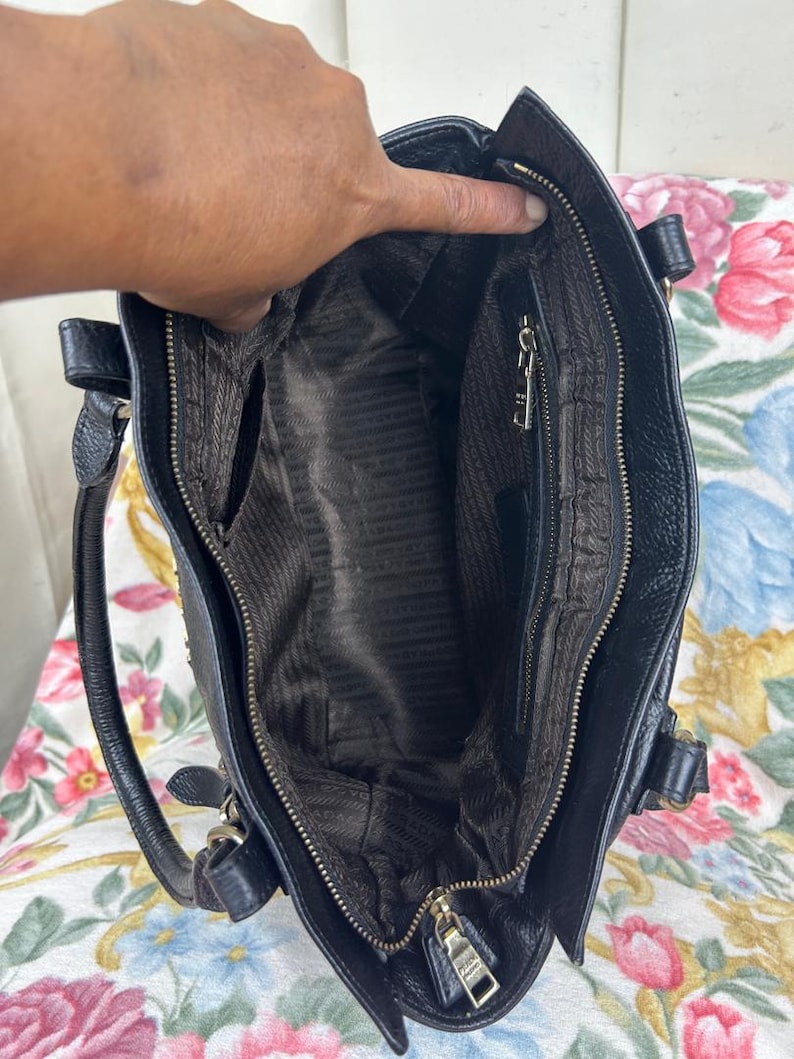 Puede incluir: Bolso de mano de cuero negro con asa superior y cierre de cremallera. El interior presenta un forro marr&oacute;n con un patr&oacute;n repetido. El bolso tiene herrajes dorados y un peque&ntilde;o bolsillo exterior.