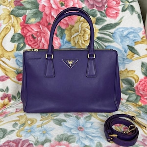 以下が含まれることがあります： ゴールドの金具が付いた濃い紫色のハンドバッグ。ファスナーと小さなロゴが付いています。バッグには2つのハンドルと取り外し可能なショルダーストラップが付いています。バッグは花柄の生地の上に置かれています。