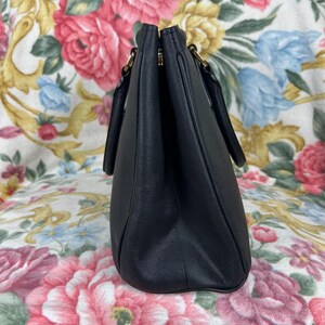 Op de afbeelding: Een zwarte handtas met twee handvatten en goudkleurige hardware. De tas is gemaakt van een glad materiaal en heeft een gestructureerde vorm. De achtergrond is een bloemenpatroon met roze, rode en blauwe bloemen.
