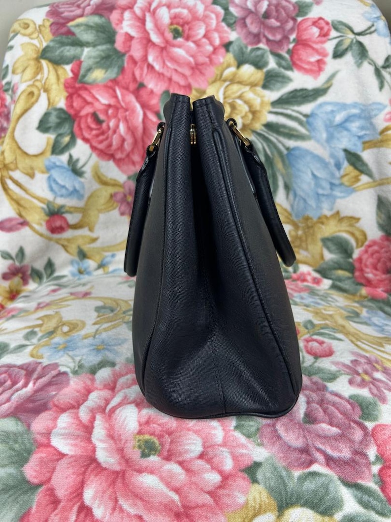 Op de afbeelding: Een zwarte handtas met twee handvatten en goudkleurige hardware. De tas is gemaakt van een glad materiaal en heeft een gestructureerde vorm. De achtergrond is een bloemenpatroon met roze, rode en blauwe bloemen.