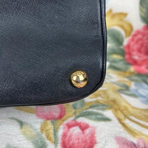 Op de afbeelding: Detail van een zwarte handtas met een getextureerd oppervlak en een goudkleurige, afgeronde stud. De hoek van de tas is zichtbaar. De achtergrond heeft een bloemenpatroon met roze en gele bloemen.