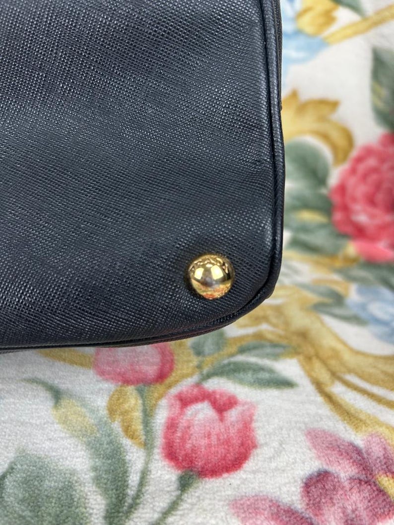Op de afbeelding: Detail van een zwarte handtas met een getextureerd oppervlak en een goudkleurige, afgeronde stud. De hoek van de tas is zichtbaar. De achtergrond heeft een bloemenpatroon met roze en gele bloemen.