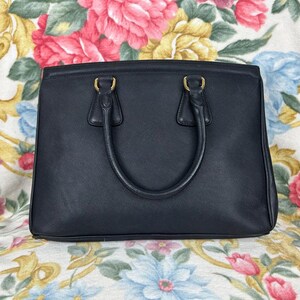 Op de afbeelding: Een zwarte leren handtas met een rechthoekige vorm en twee handvatten. De tas heeft goudkleurige hardware en een gestructureerd ontwerp. De achtergrond heeft een bloemenpatroon met roze, gele en blauwe bloemen.
