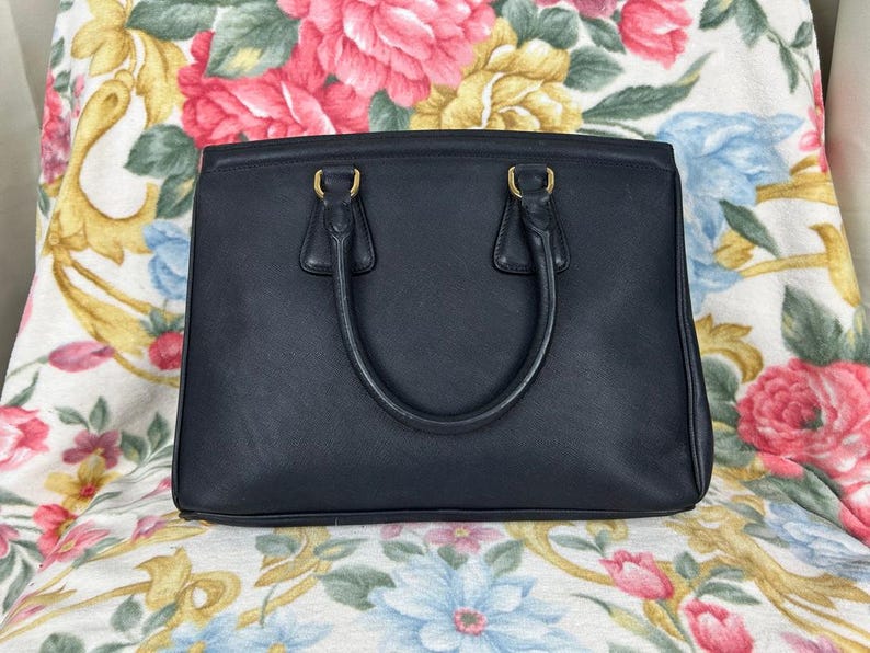 Op de afbeelding: Een zwarte leren handtas met een rechthoekige vorm en twee handvatten. De tas heeft goudkleurige hardware en een gestructureerd ontwerp. De achtergrond heeft een bloemenpatroon met roze, gele en blauwe bloemen.