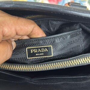 Op de afbeelding: Zwarte handtas met een gouden rits en een gouden Prada Milano logo. De binnenkant van de tas is zwart met een getextureerde voering. De tas is open en onthult de binnenkant.
