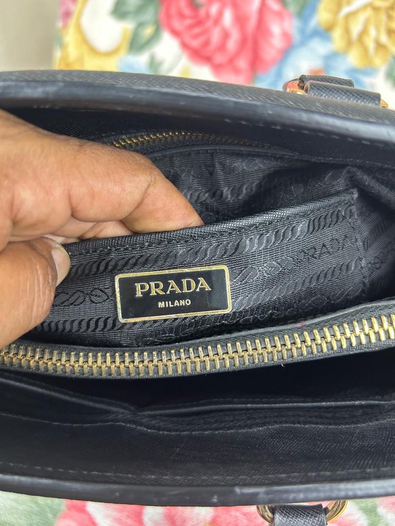 Op de afbeelding: Zwarte handtas met een gouden rits en een gouden Prada Milano logo. De binnenkant van de tas is zwart met een getextureerde voering. De tas is open en onthult de binnenkant.
