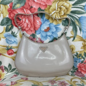 May include: A light beige Prada handbag with a curved shape and a short handle. The bag has a shiny, patent leather finish and a small Prada logo on the front. The background is a floral pattern with pink, blue, and yellow flowers.