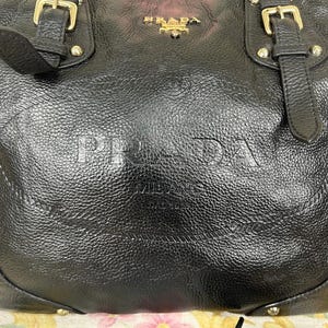 Puede incluir: Bolso de mano de cuero negro con letras en relieve "PRADA MILANO". El bolso presenta herrajes dorados, incluyendo hebillas y tachuelas. La parte superior del bolso tiene el logotipo dorado "PRADA". El cuero tiene una apariencia texturizada.
