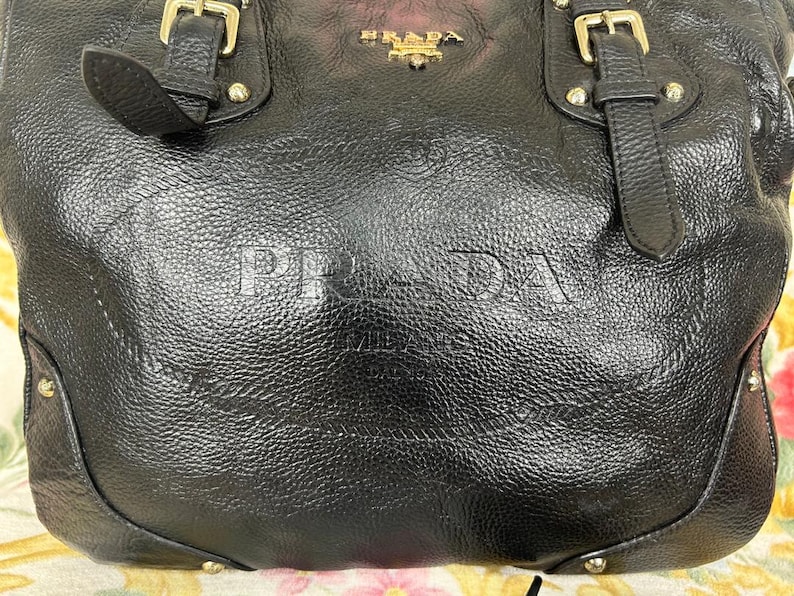 Puede incluir: Bolso de mano de cuero negro con letras en relieve "PRADA MILANO". El bolso presenta herrajes dorados, incluyendo hebillas y tachuelas. La parte superior del bolso tiene el logotipo dorado "PRADA". El cuero tiene una apariencia texturizada.