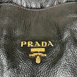 Puede incluir: Primer plano de un bolso de mano de cuero negro con el logotipo dorado "PRADA MILANO" en un lugar destacado. El cuero tiene un aspecto texturizado y granulado. El dise&ntilde;o del bolso sugiere un accesorio de moda de lujo.