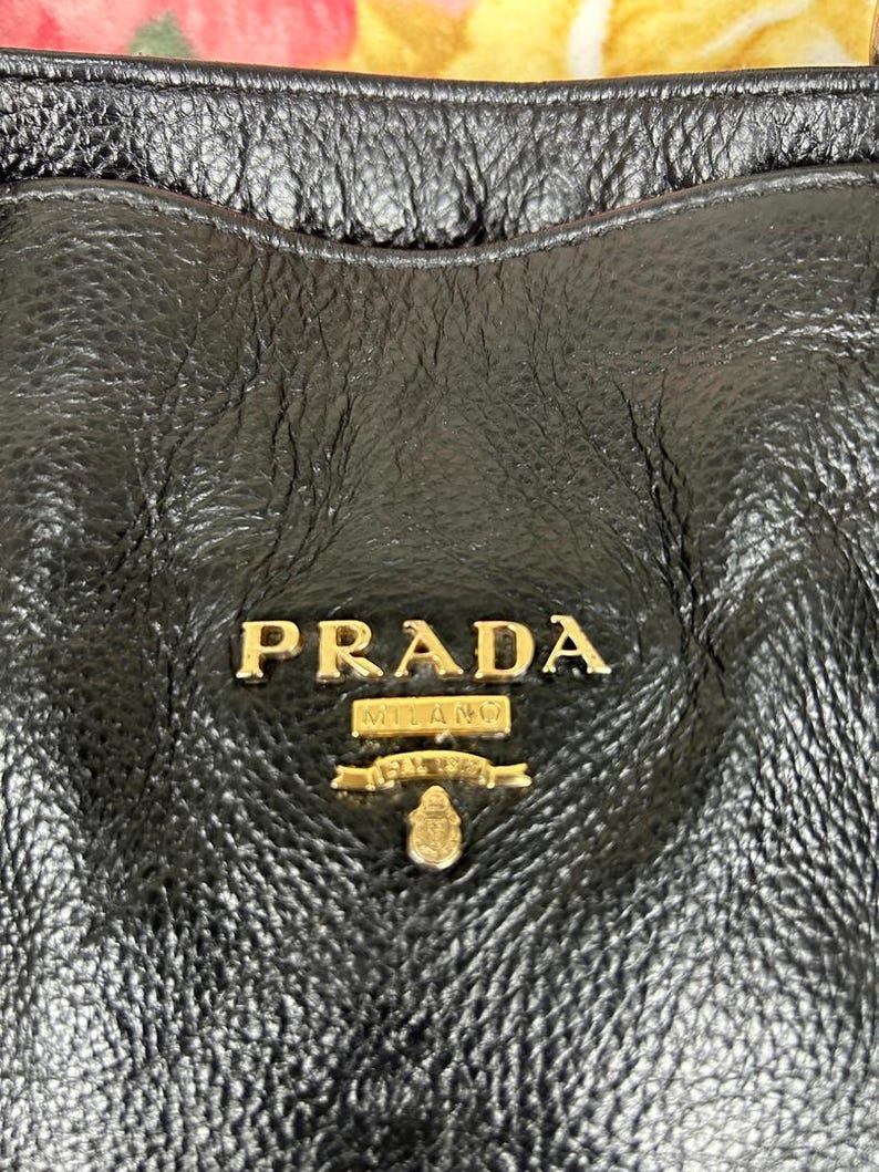 Puede incluir: Primer plano de un bolso de mano de cuero negro con el logotipo dorado "PRADA MILANO" en un lugar destacado. El cuero tiene un aspecto texturizado y granulado. El dise&ntilde;o del bolso sugiere un accesorio de moda de lujo.