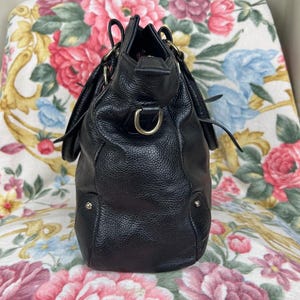 Puede incluir: Un bolso de mano de cuero negro con un acabado texturizado. El bolso presenta herrajes dorados, incluyendo una anilla en D y peque&ntilde;os tachuelas. Tiene una forma estructurada con un asa superior y una fijaci&oacute;n para correa de hombro.