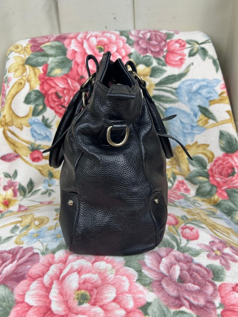 Puede incluir: Un bolso de mano de cuero negro con un acabado texturizado. El bolso presenta herrajes dorados, incluyendo una anilla en D y peque&ntilde;os tachuelas. Tiene una forma estructurada con un asa superior y una fijaci&oacute;n para correa de hombro.