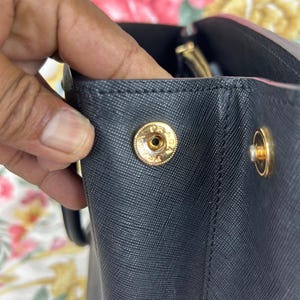 Op de afbeelding: Close-up van een zwarte handtas met goudkleurige hardware. De tas heeft een getextureerd oppervlak en een druksluiting met de letters "PR" gegraveerd op de knop. De handgreep van de tas is zichtbaar op de achtergrond.