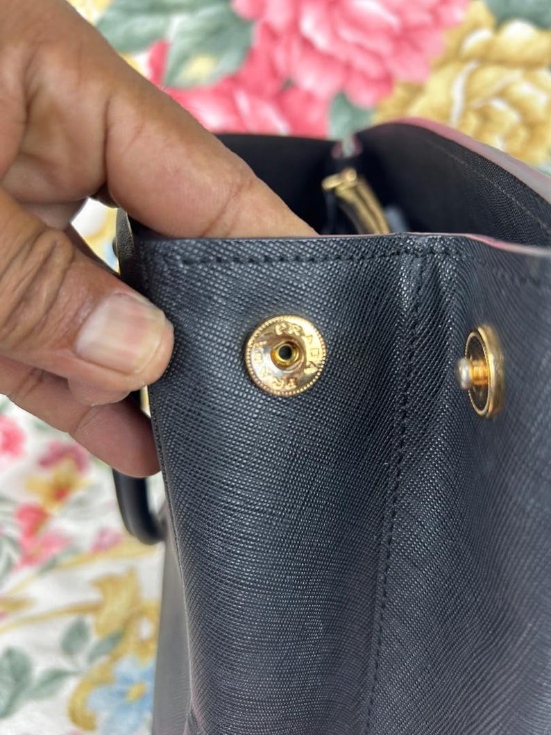 Op de afbeelding: Close-up van een zwarte handtas met goudkleurige hardware. De tas heeft een getextureerd oppervlak en een druksluiting met de letters "PR" gegraveerd op de knop. De handgreep van de tas is zichtbaar op de achtergrond.