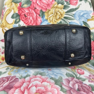 Puede incluir: Bolso de mano de cuero negro con un acabado texturizado. La parte inferior del bolso presenta cuatro patas met&aacute;licas y un dise&ntilde;o estructurado. El bolso est&aacute; sobre un fondo con estampado floral.