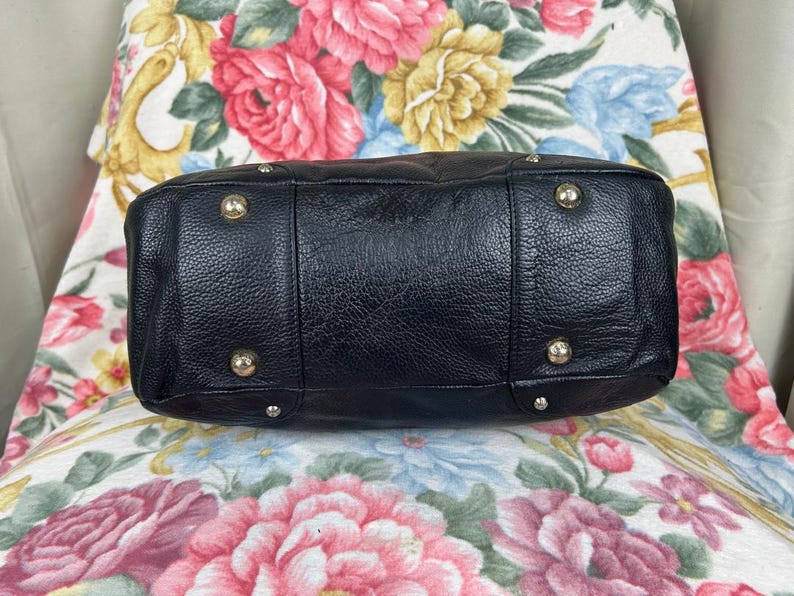 Puede incluir: Bolso de mano de cuero negro con un acabado texturizado. La parte inferior del bolso presenta cuatro patas met&aacute;licas y un dise&ntilde;o estructurado. El bolso est&aacute; sobre un fondo con estampado floral.