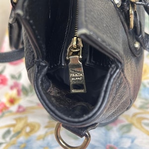 Puede incluir: Primer plano de un bolso de mano de cuero negro con cremallera y herrajes dorados. El tirador de la cremallera lleva grabado "PRADA MILANO". El bolso tiene una forma estructurada y un asa superior. El interior es visible.