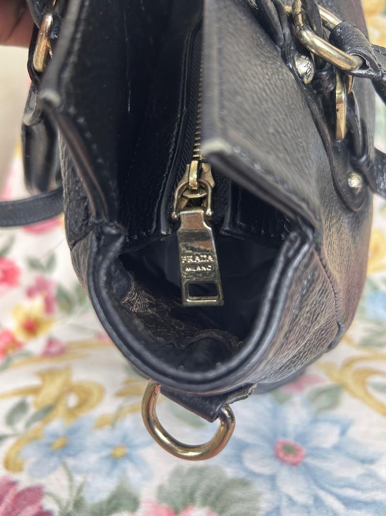 Puede incluir: Primer plano de un bolso de mano de cuero negro con cremallera y herrajes dorados. El tirador de la cremallera lleva grabado "PRADA MILANO". El bolso tiene una forma estructurada y un asa superior. El interior es visible.
