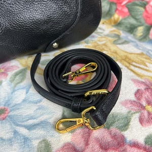 Puede incluir: Bolso de mano de cuero negro con una correa de hombro desmontable. La correa est&aacute; enrollada y presenta cierres met&aacute;licos dorados. El bolso tiene una superficie texturizada y herrajes dorados.