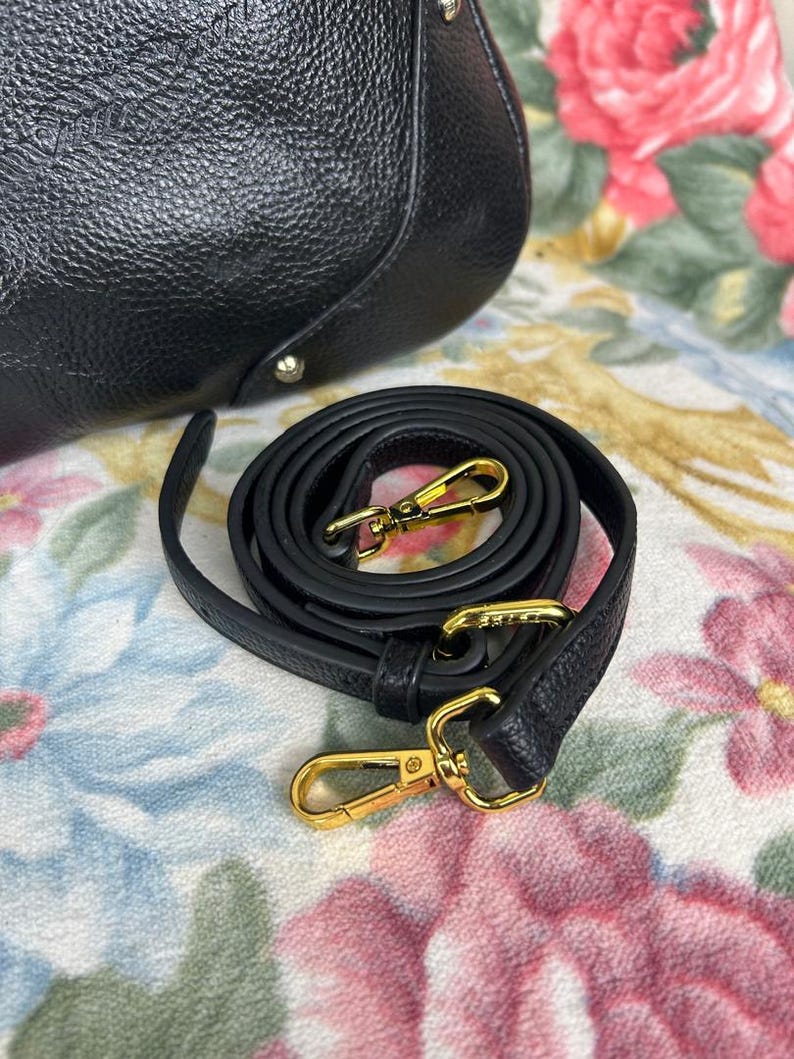 Puede incluir: Bolso de mano de cuero negro con una correa de hombro desmontable. La correa est&aacute; enrollada y presenta cierres met&aacute;licos dorados. El bolso tiene una superficie texturizada y herrajes dorados.