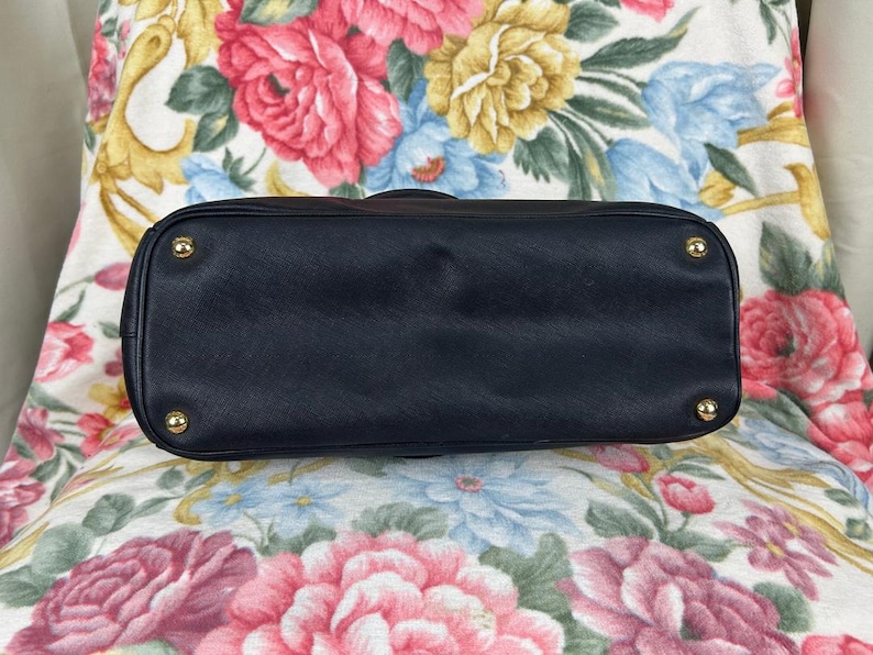 Op de afbeelding: Onderkant van een marineblauwe handtas. De tas heeft een rechthoekige vorm met afgeronde hoeken. Goudkleurige metalen studs zijn zichtbaar op de hoeken. De tas is gemaakt van een glad, effen materiaal.